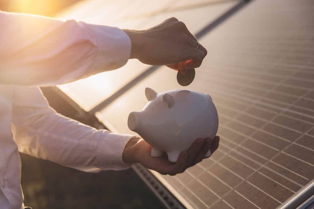 Person saving money with piggy bank near solar panels at sunset.