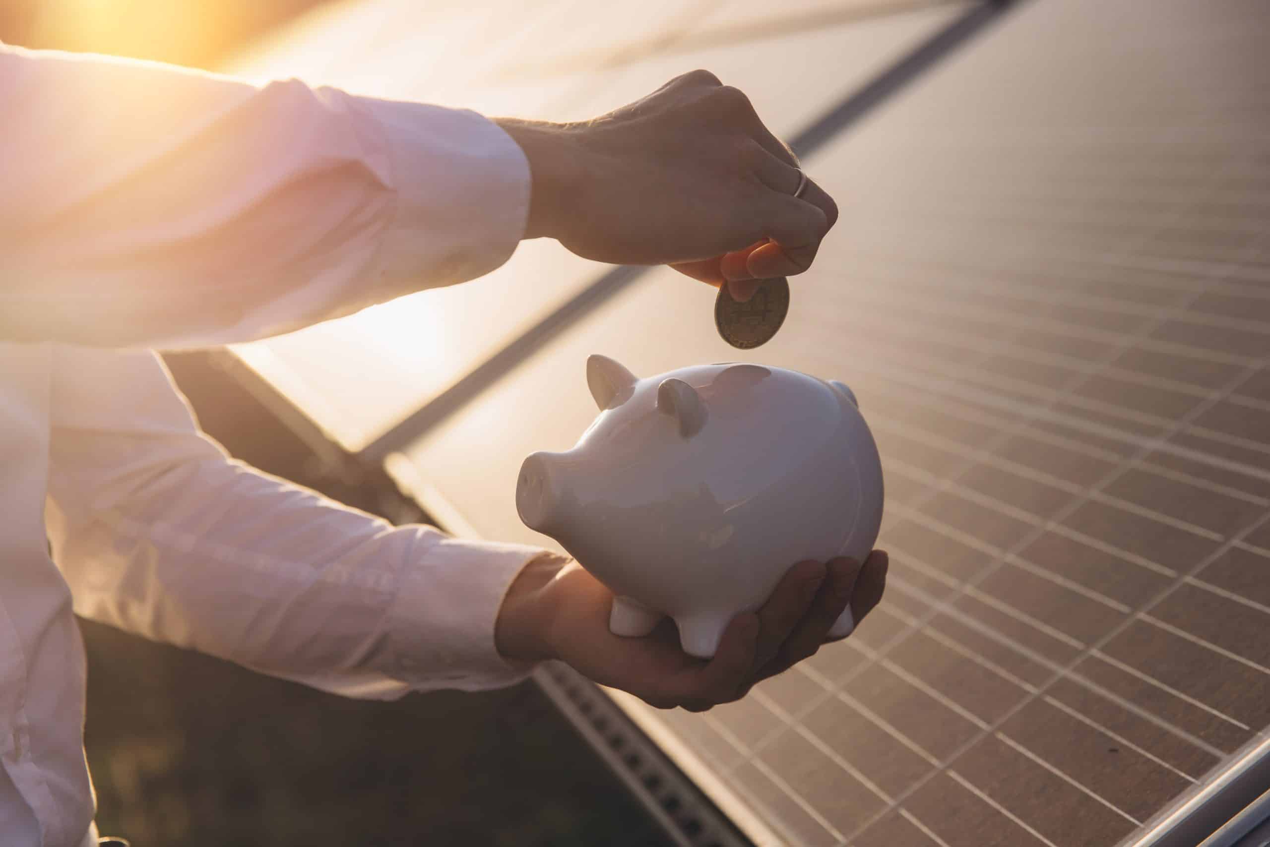 Person saving money with piggy bank near solar panels at sunset.
