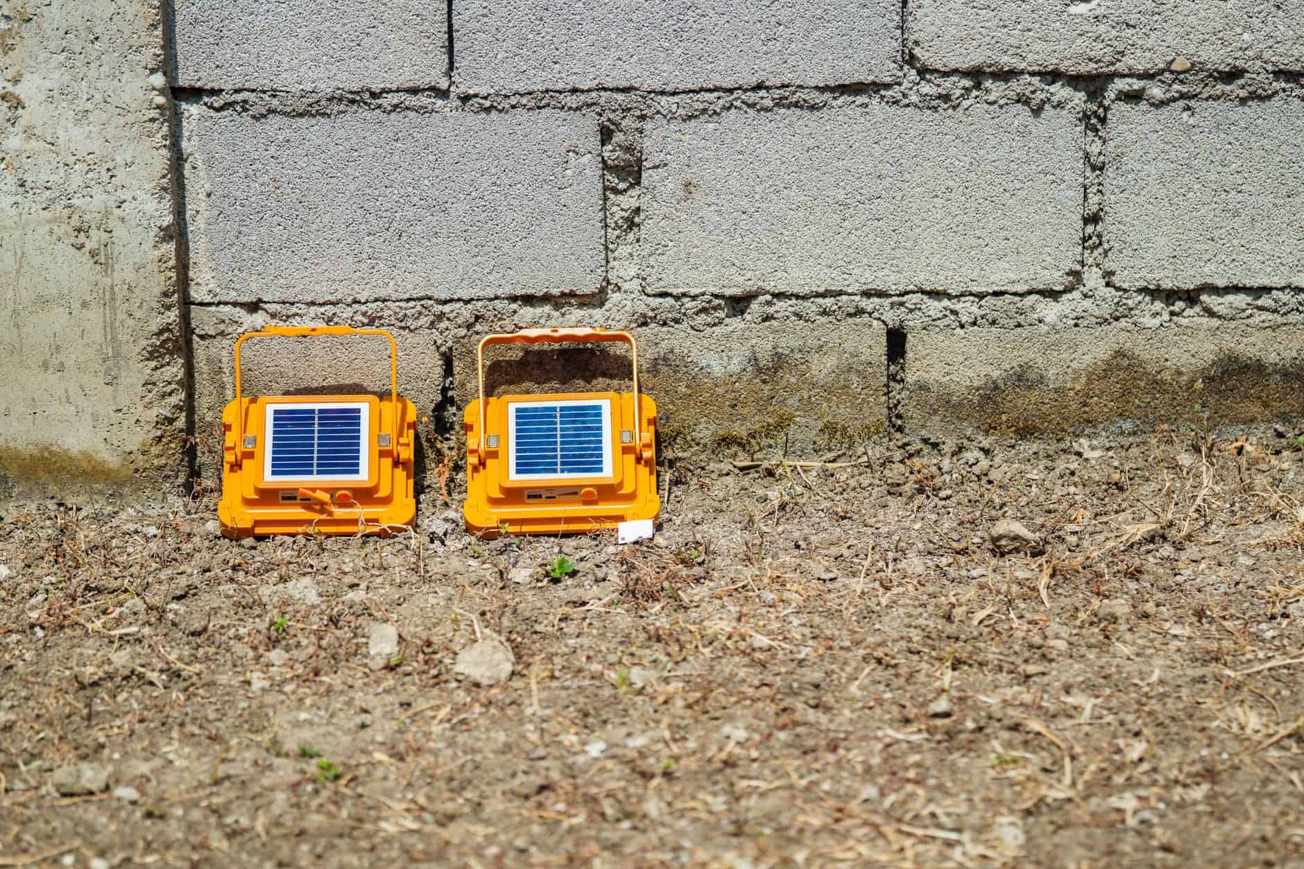 Two small solar-powered devices placed on the ground near a brick wall.