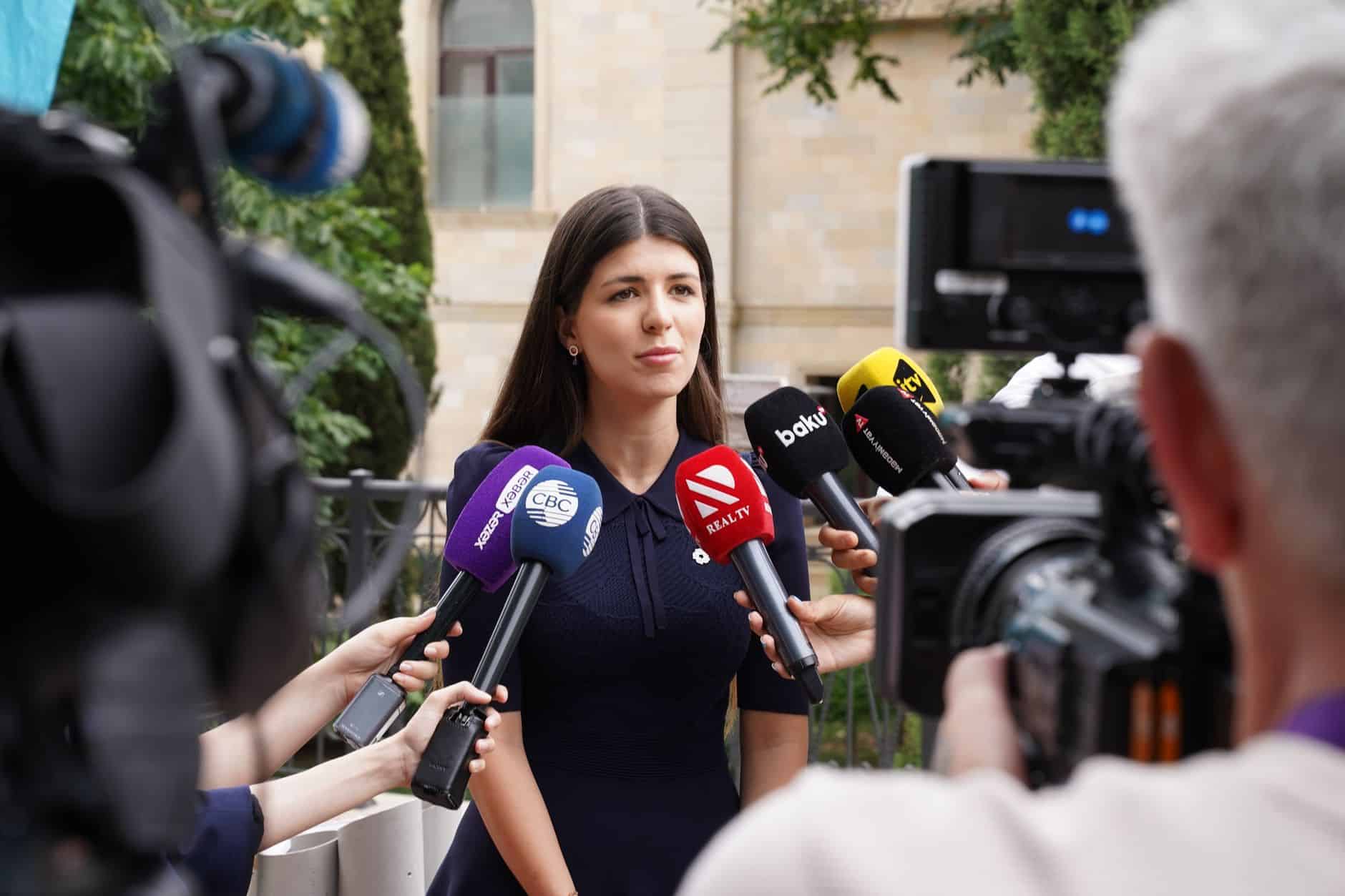 A spokesperson engages with media during an outdoor press interview surrounded by microphones.