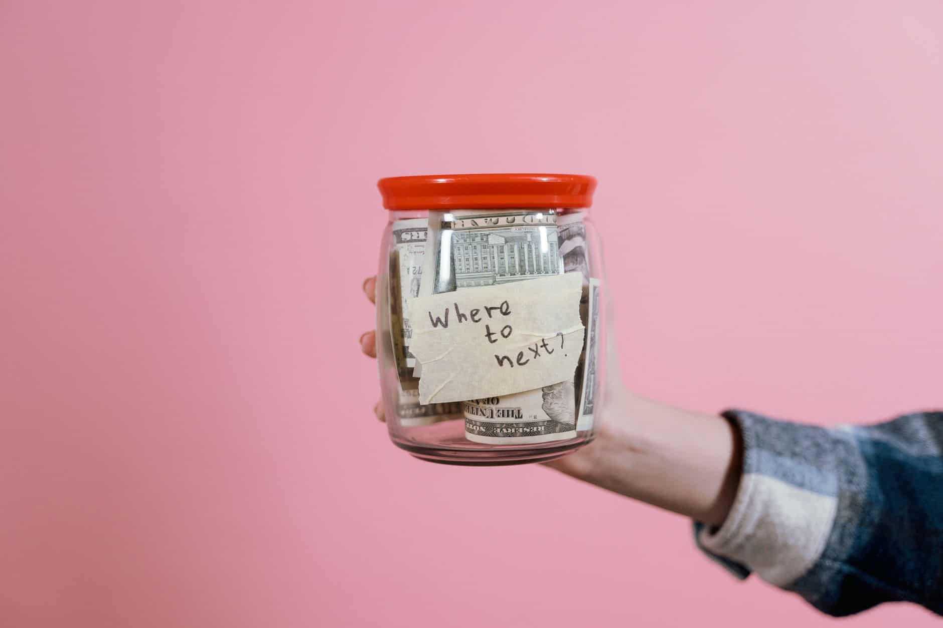 Een hand houdt een pot vast gevuld met geld en een briefje met de tekst 'Waar gaan we nu naartoe?'.