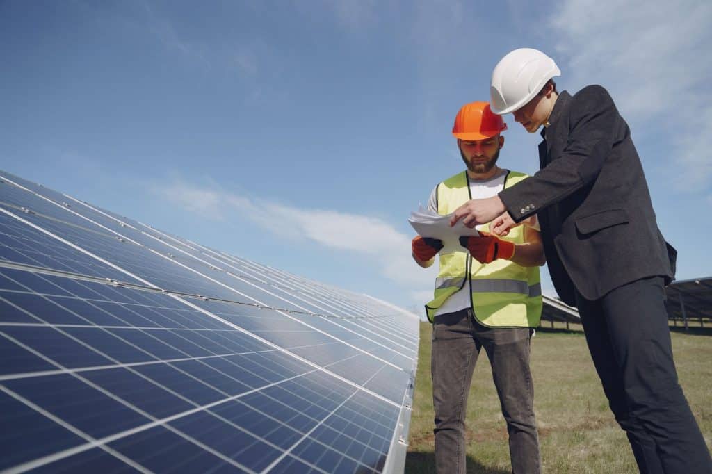 Low angle of young inspector and foreman in hardhats checking documentation against modern solar panels in field