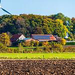 Solar farm with wind turbine and farmhouse under blue sky.