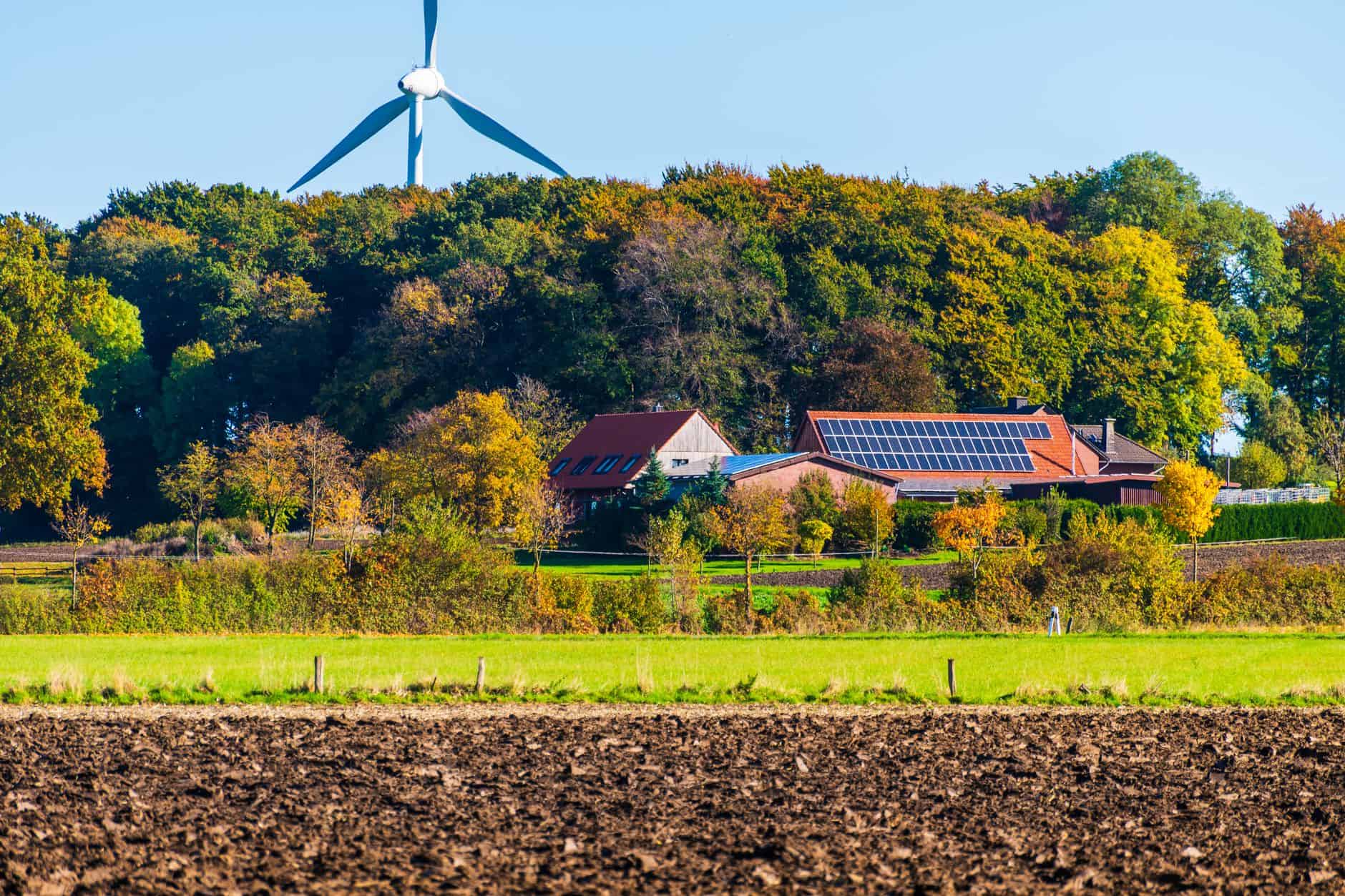 Solar farm with wind turbine and farmhouse under blue sky.