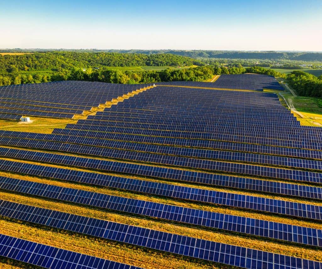 Large solar panel farm in a rural landscape with green fields and trees in the background.