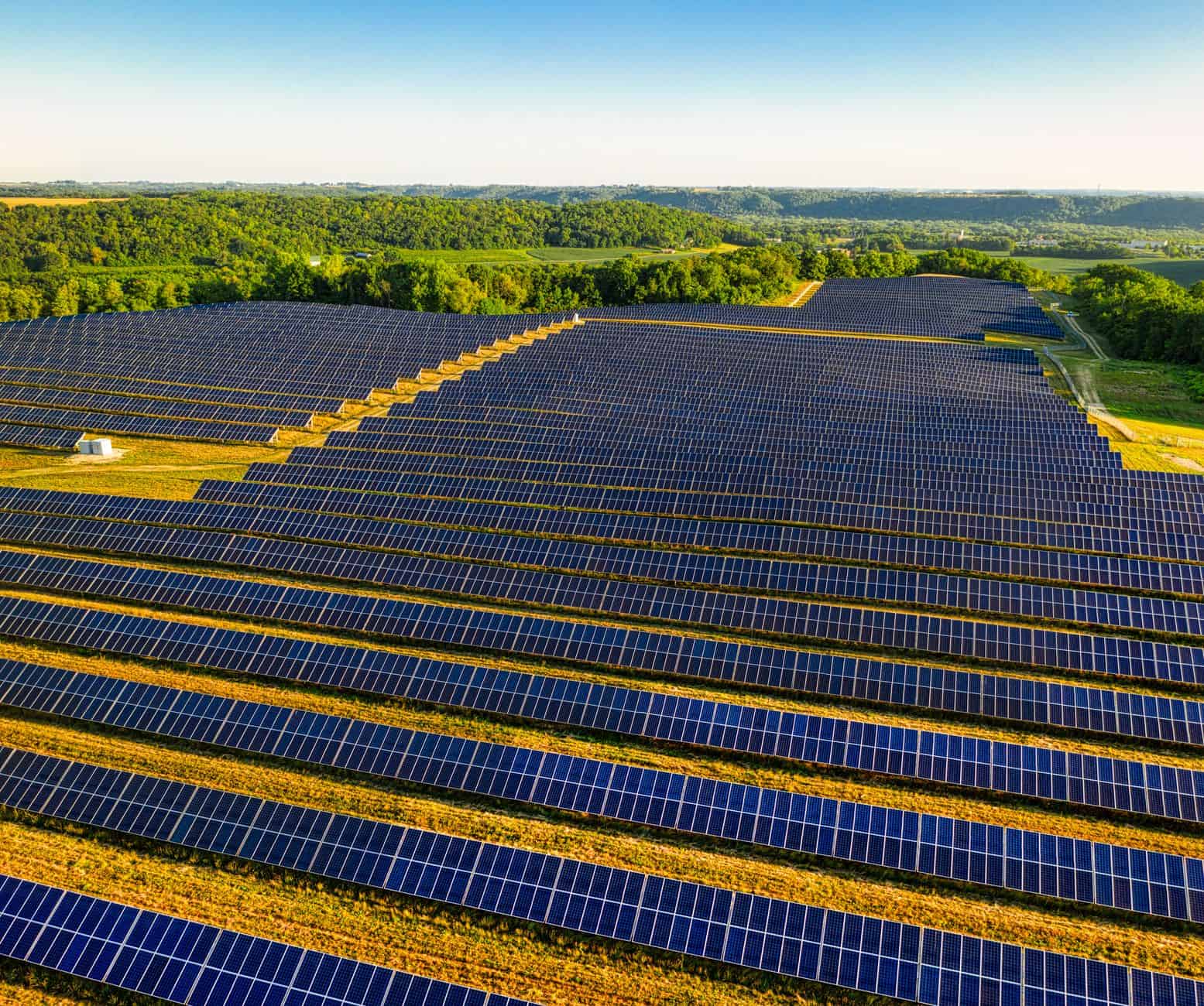 Large solar panel farm in a rural landscape with green fields and trees in the background.
