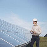 Young serious male engineer in formal outfit and helmet reporting while inspecting solar panel field