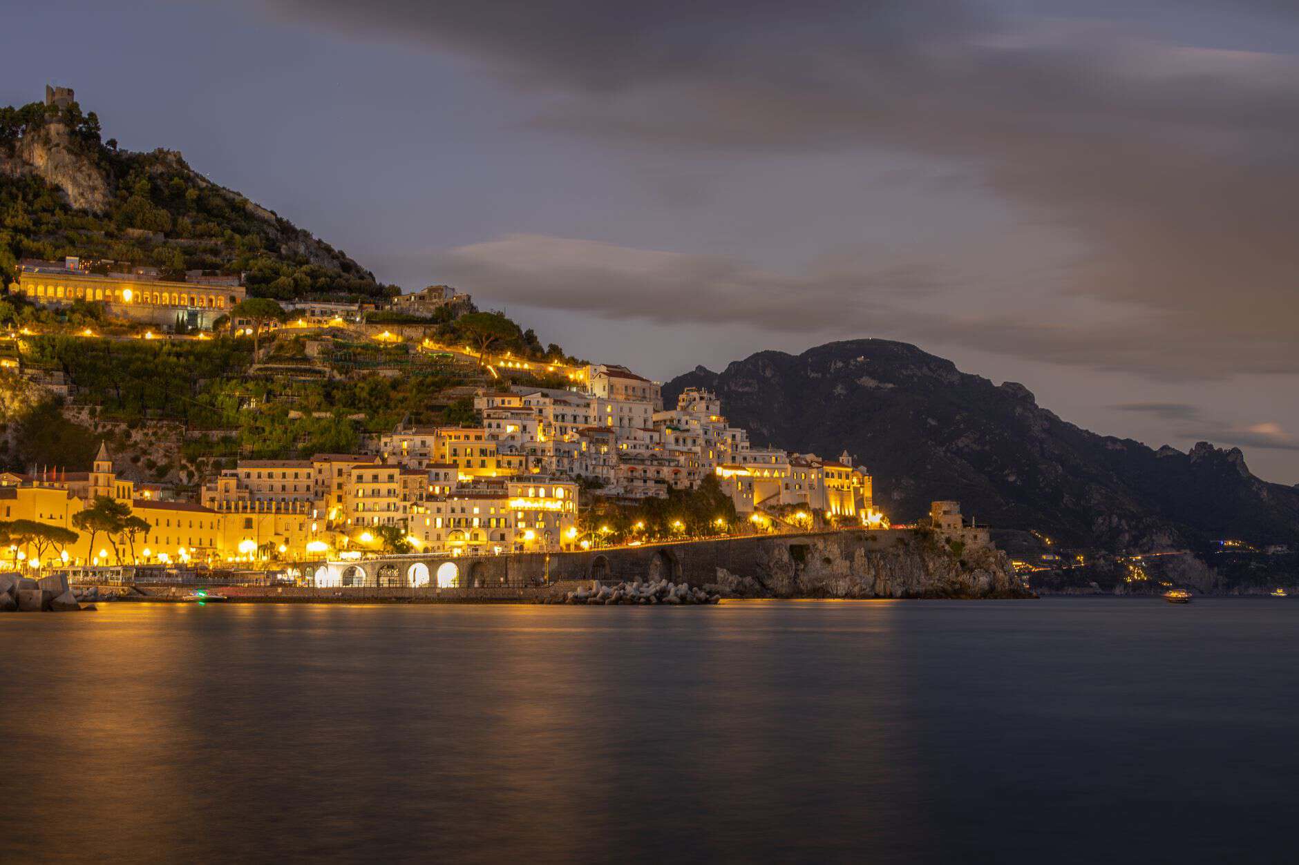 Night view of a coastal town with illuminated buildings and dark mountains in the background.